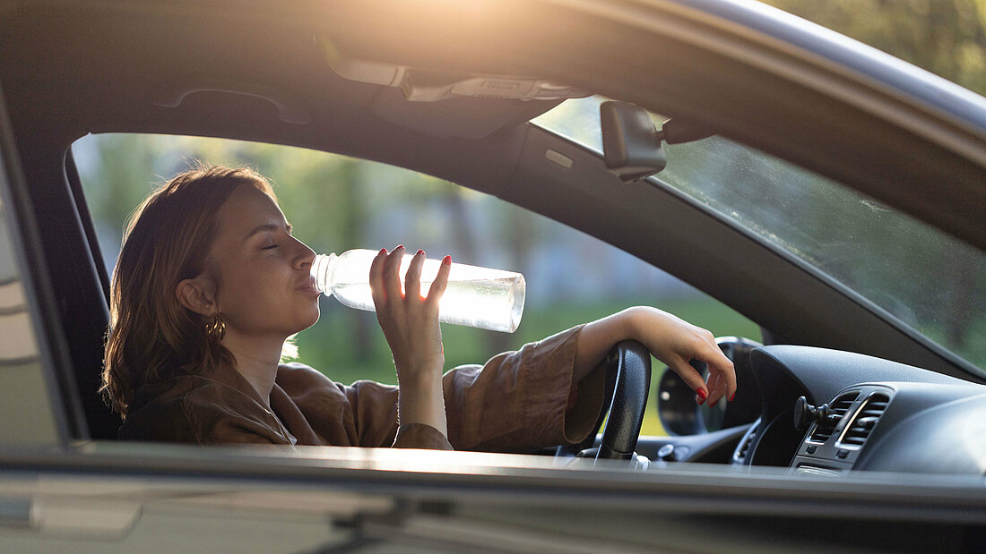 A woman sits at the wheel of a car and takes a sip of water from a bottle.