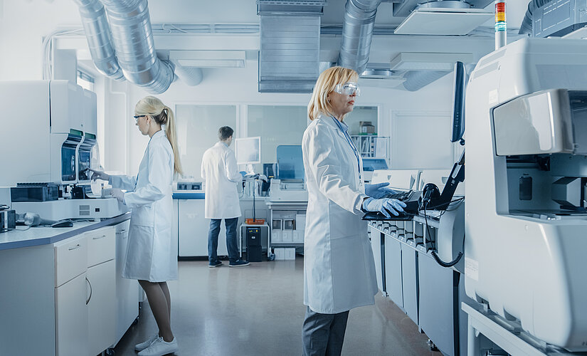 Two women and a man wearing white coats and protective goggles stand in a laboratory.