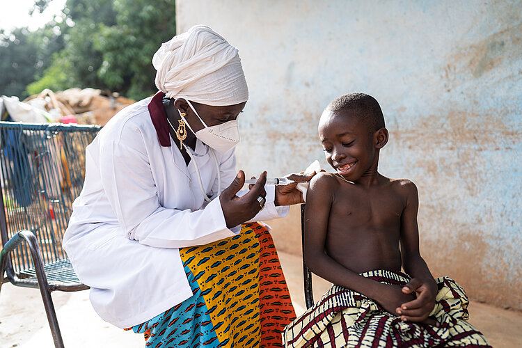 A woman in a white coat sticks a syringe into the arm of a laughing child.