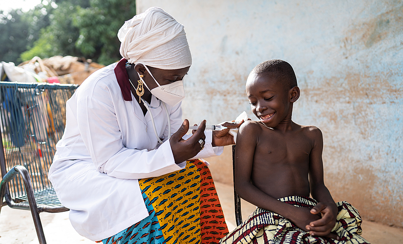 A woman in a white coat sticks a syringe into the arm of a laughing child.