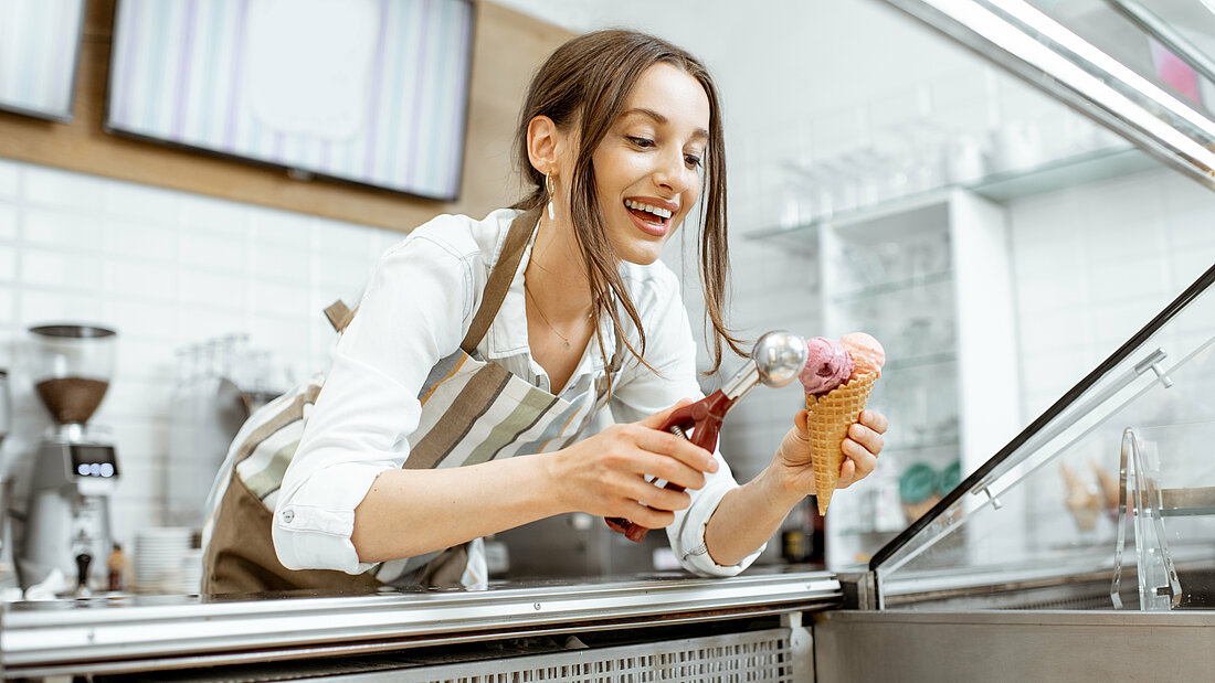 A laughing young woman holds an ice cream scoop in one hand and a wafer cone with two scoops of ice cream in the other.