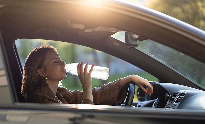 A woman sits at the wheel of a car and takes a sip of water from a bottle.